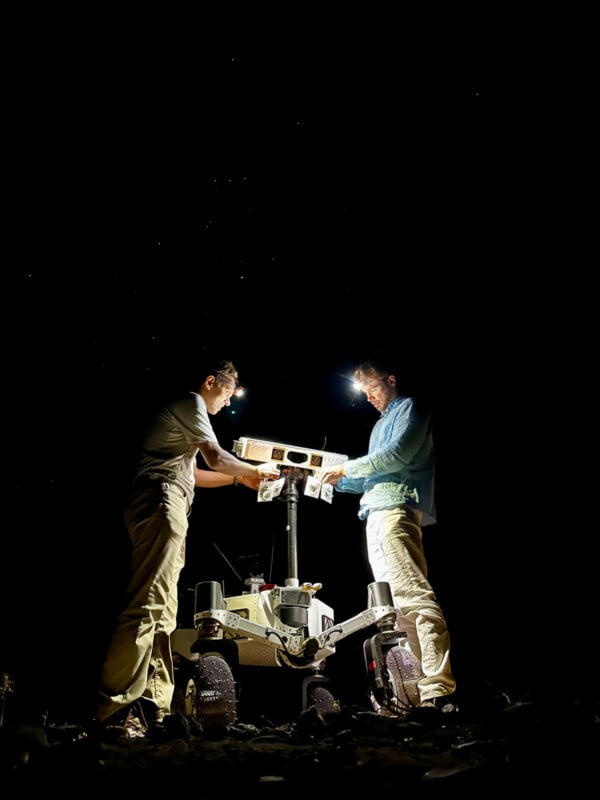Two people wearing headlamps work at night on a rover-like robotic vehicle under a starry sky, illuminated by their lights.