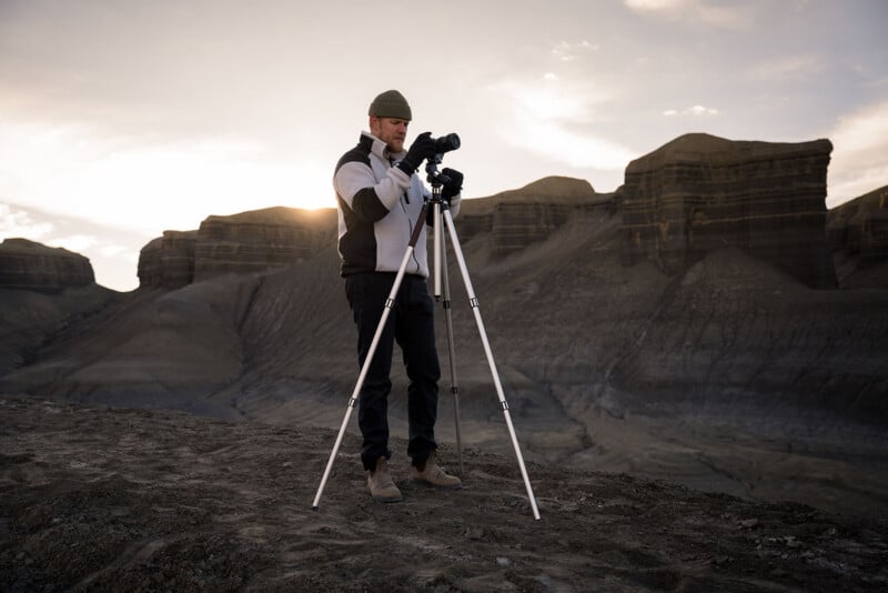 A person in outdoor clothing adjusts a camera on a tripod, standing on rocky ground with large, layered cliffs and soft sunlight in the background.