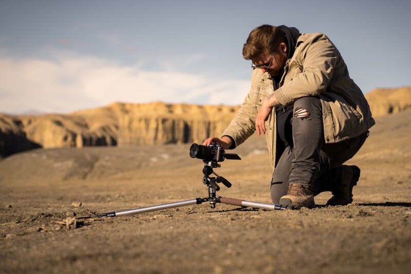 A man kneels on rocky ground in a desert landscape, adjusting a camera mounted on a low tripod, with tan cliffs visible in the background under a clear sky.