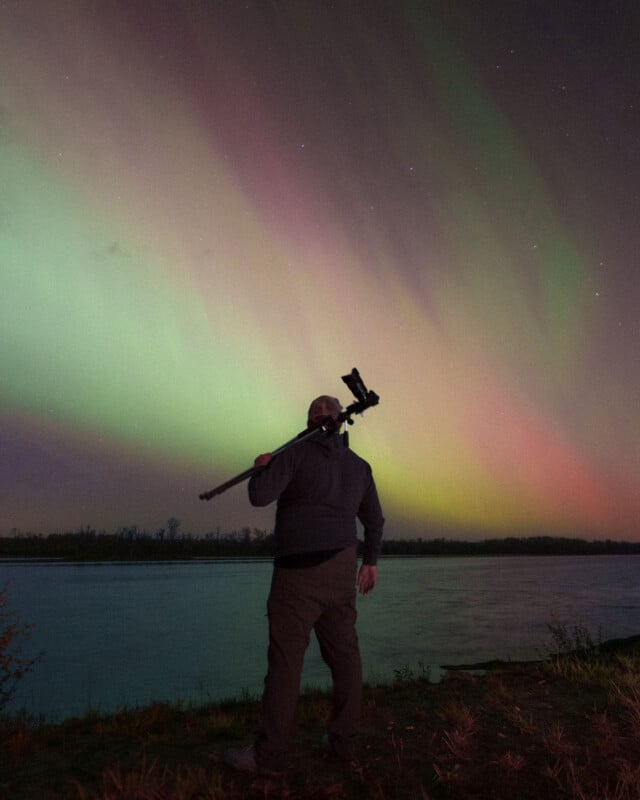 A person stands by a riverbank at night, holding a tripod over their shoulder, watching vibrant green and pink aurora lights dancing across the sky.