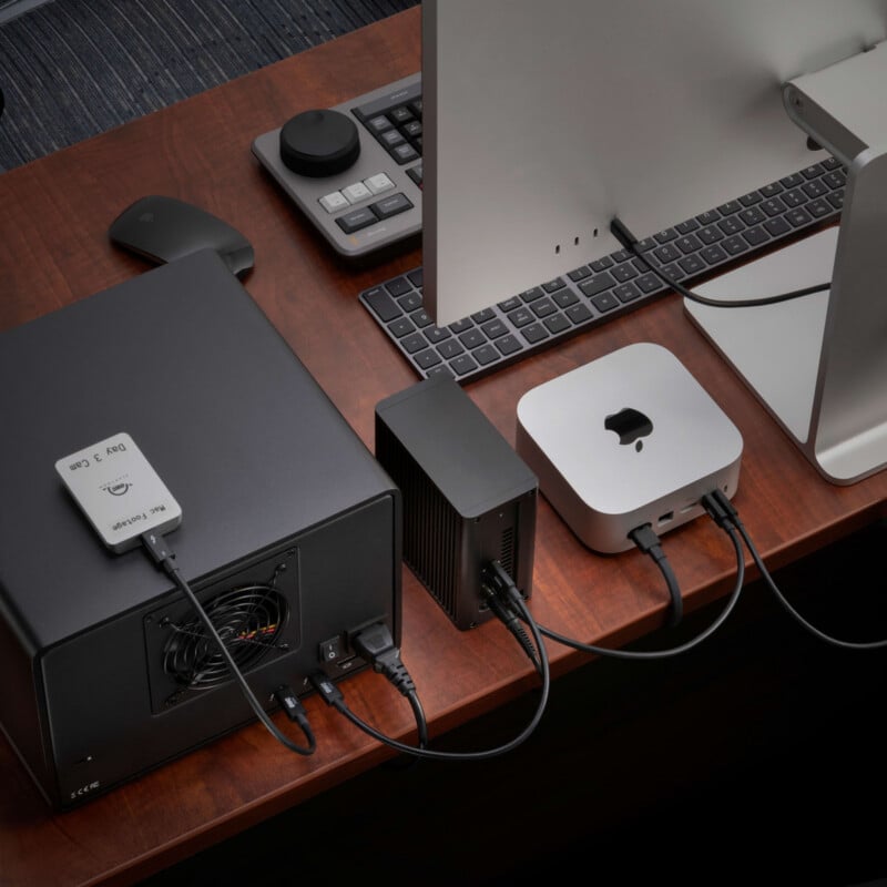 A desktop computer, external drive, hub, and small device are connected with cables on a wooden desk, along with a keyboard and control pad.