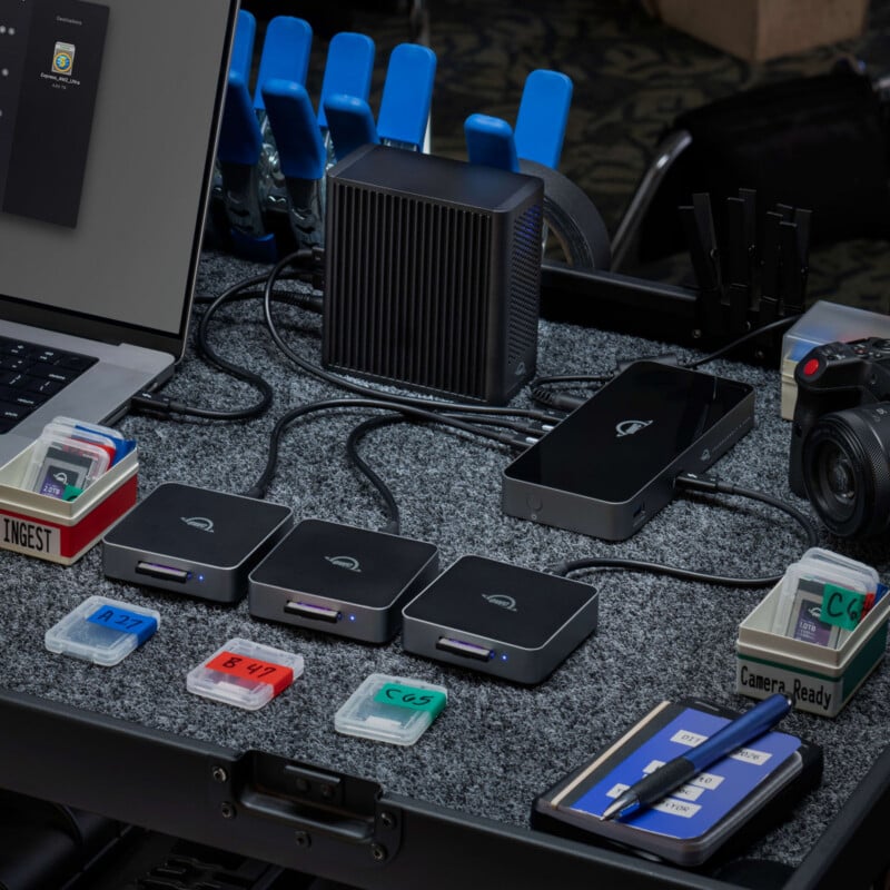 A workstation with a laptop, card readers, memory cards, a digital camera, a black device, cables, and labeled trays on a gray felt surface. Blue and black clamps are visible in the background.