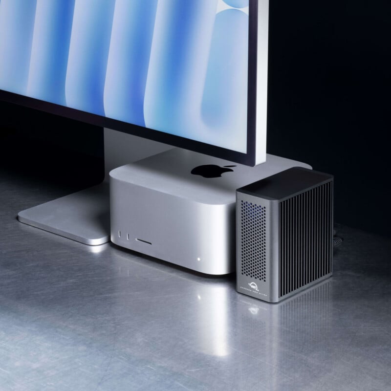 A silver Apple Mac Studio, a black external drive, and a monitor with a blue abstract design sit on a reflective metallic surface.