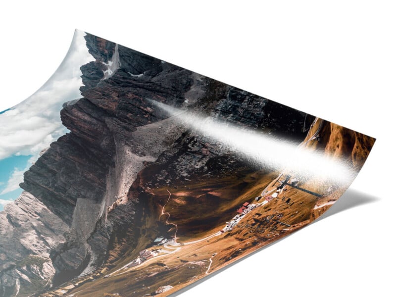 A glossy poster print of a mountainous landscape, featuring rocky peaks, winding roads, and scattered buildings, with light reflecting off the image’s surface.