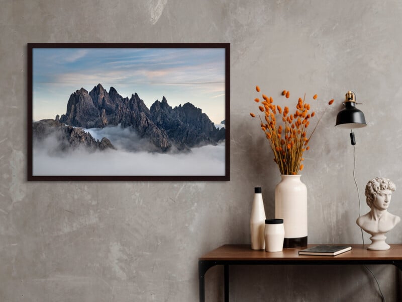 A framed mountain landscape photo hangs on a gray wall above a wooden table with vases, an orange floral arrangement, a black lamp, and a classical bust sculpture.