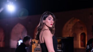 A woman with long hair and red lipstick stands in a black dress, looking over her shoulder at the camera. Behind her, a person plays piano under dramatic lighting, with arched structures in the background.