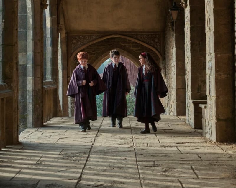 Three children in robes walk down a stone corridor with archways, talking and smiling. Sunlight casts shadows on the floor. The scene looks old-fashioned and magical.