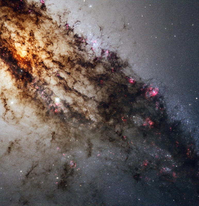 A vibrant galaxy with bright stars, glowing orange and pink regions, and dense dark dust lanes stretching diagonally across a star-filled background in space.