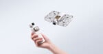 A hand holds a white handheld camera with a small screen, while a white drone with a protective cage hovers in the air.