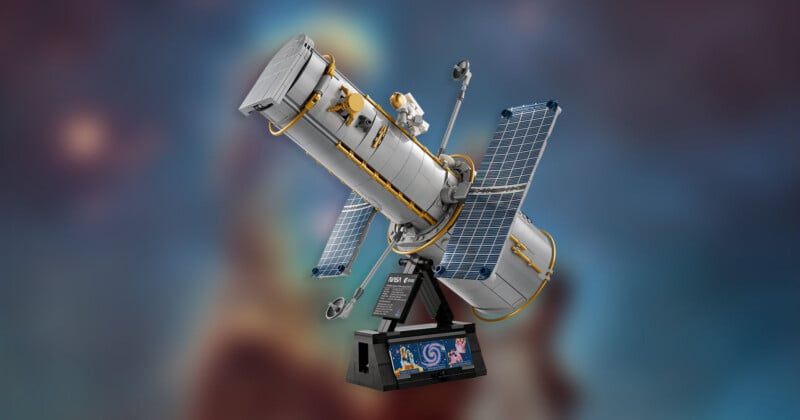 LEGO model of the Hubble Space Telescope with solar panels and mini astronaut figure, displayed on a black stand with a galaxy image, against a blurred cosmic background.