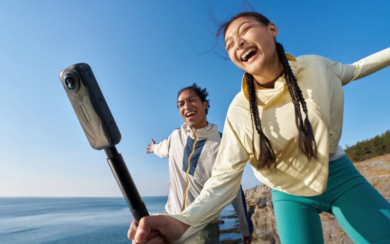 Two people smile and pose energetically on a seaside cliff, holding a 360-degree camera on a selfie stick, with the blue sky and ocean in the background.