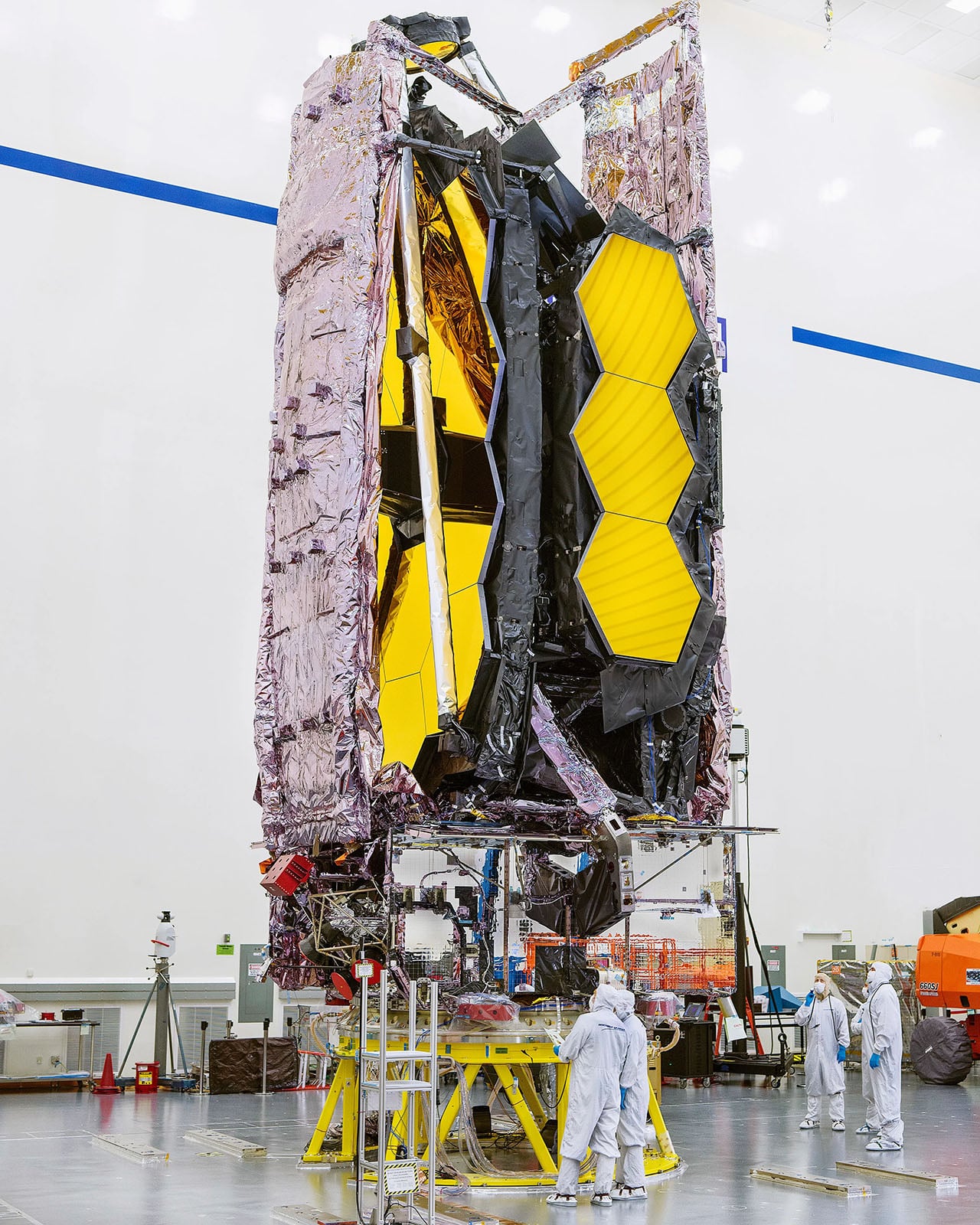 Engineers in cleanroom suits work near the large, gold-paneled James Webb Space Telescope, which stands upright with reflective foil and hexagonal mirrors visible inside a bright, spacious laboratory.