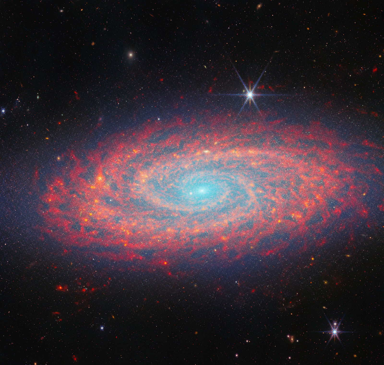 A colorful spiral galaxy glows with blue, pink, and red hues against a dark star-filled sky. Bright stars with diffraction spikes appear in the foreground.