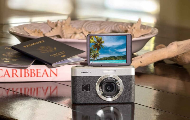 A digital camera with a flip-up screen shows a tropical beach. Behind it are passports, a "Caribbean" guidebook, seashells in a bowl, and driftwood on a reflective wooden surface.