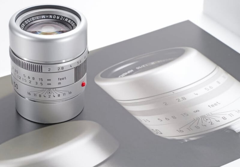 A silver camera lens sits on top of an open booklet that displays a photo of the same lens. The lens shows engraved numbers and a red dot for mounting alignment.