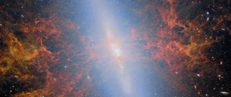 A bright spiral galaxy surrounded by colorful red and orange interstellar clouds, set against a backdrop of dark space dotted with stars.