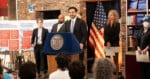 A man speaks at a podium with a city seal, flanked by officials. Behind them are American flags, posters with text, and a brick wall. People in the audience listen attentively.