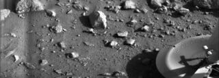 Black and white image of a rocky, dusty Martian surface with scattered stones and a spacecraft part, possibly a lander footpad, visible in the lower right corner.