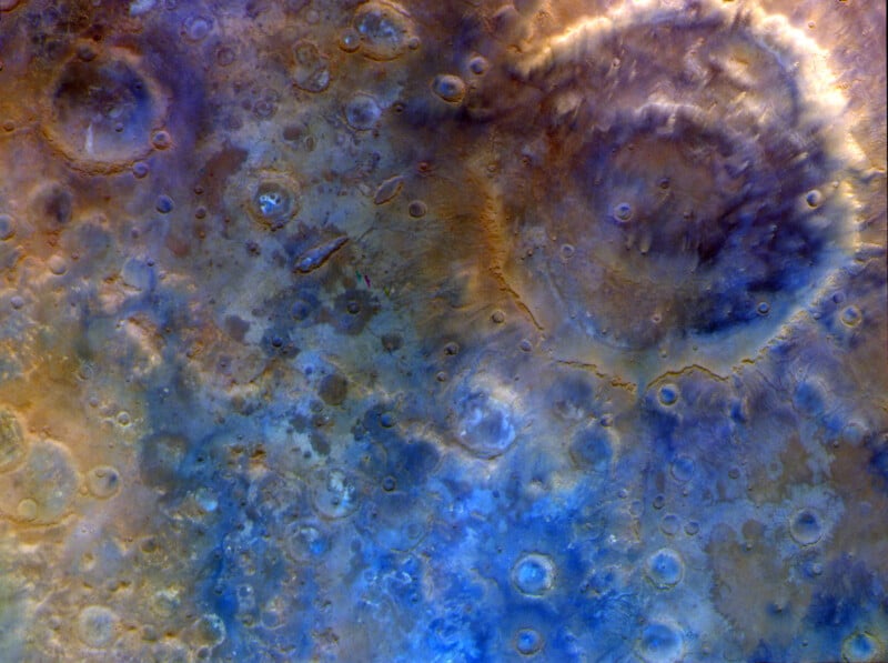 Satellite view of a planet’s surface with craters in shades of brown, blue, and purple, showing rugged terrain and circular impact marks.