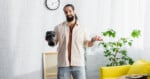 A man with a beard holds a camera in one hand and shrugs with the other, standing in a bright room with a yellow couch, green plant, and a clock on the wall.