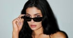 A woman with long dark hair and nude lipstick looks forward, lifting her black sunglasses with one hand against a plain light background.
