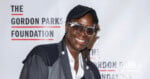A smiling person in glasses and a dark cap poses in front of a backdrop that reads