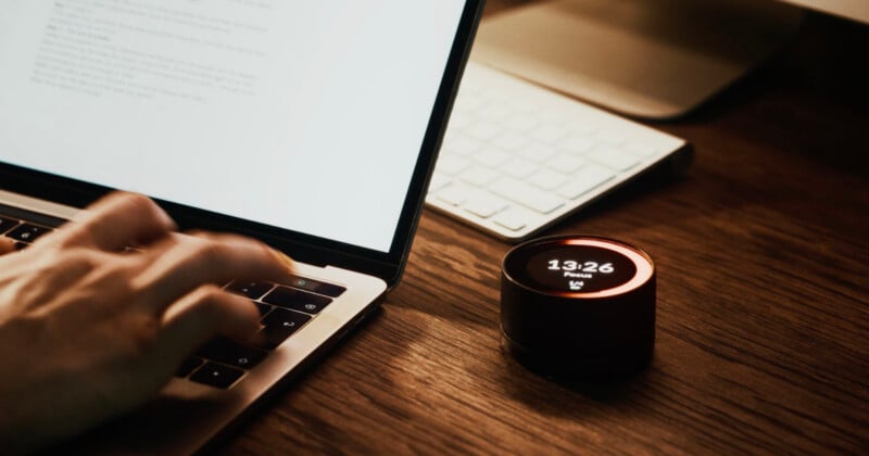 A person types on a laptop at a wooden desk. Next to the laptop is a small round digital clock displaying "13:26" and "Pause" with a glowing red ring. A wireless keyboard is in the background.