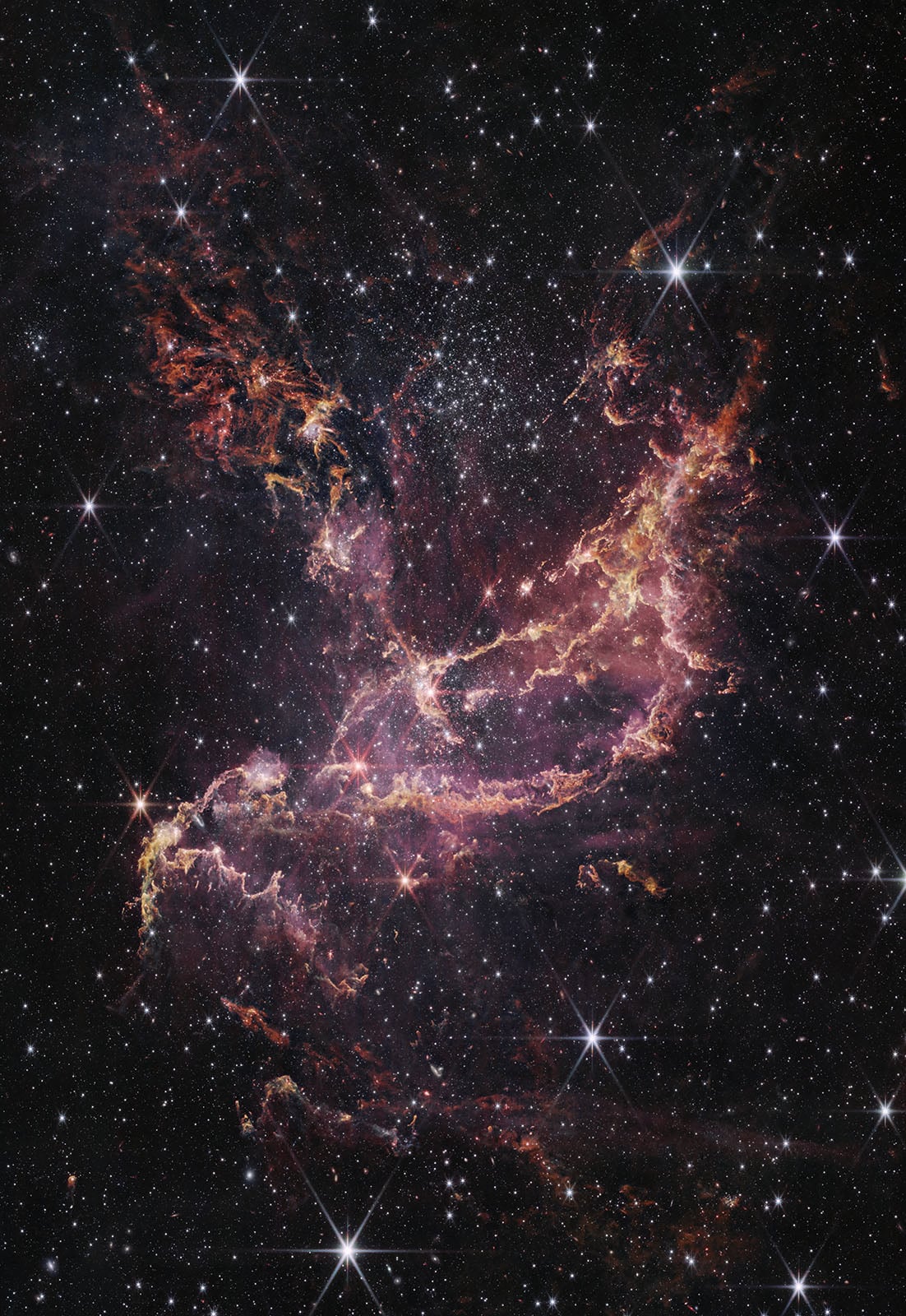 A vibrant nebula glows with pink, red, and yellow clouds of gas and dust, surrounded by countless bright stars scattered across the dark expanse of space.