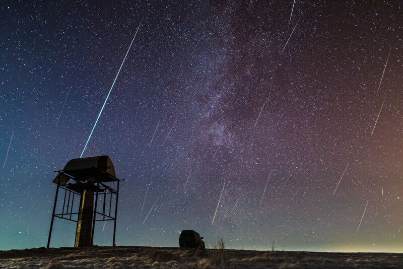 A night sky filled with stars and numerous meteor streaks, with a silhouetted metal observation structure and small building on the ground beneath the celestial display.