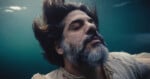 A bearded man with closed eyes and flowing hair floats underwater, wearing a tattered light shirt, appearing serene and calm.