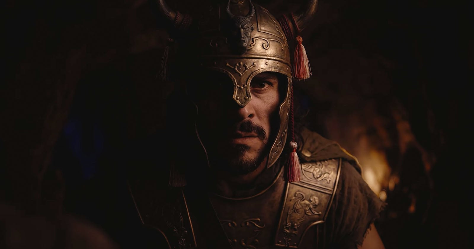 A man wearing a horned, bronze helmet and ornate armor stares intensely forward in a dimly lit setting, with shadows highlighting his serious expression.