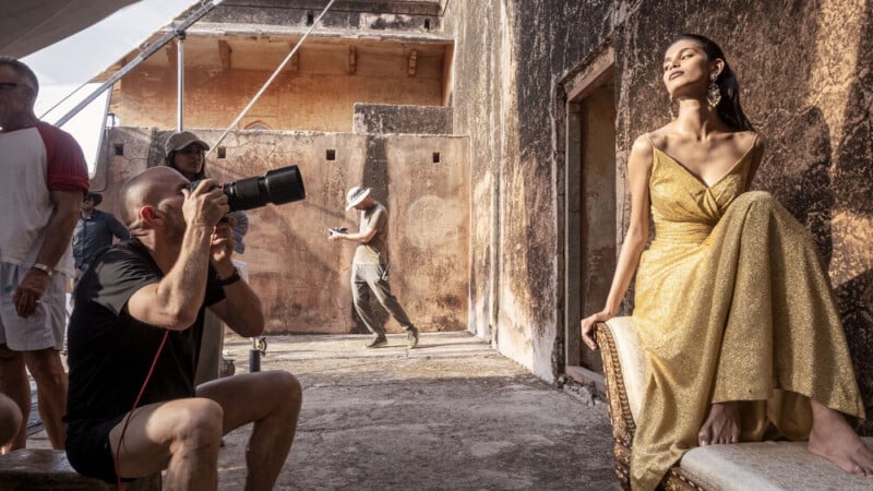A woman in a gold dress poses on a chair against a weathered wall as a photographer takes her picture. Several people stand and walk in the sunlit, rustic setting.