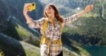 A smiling woman with wavy hair and a yellow backpack wearing a plaid shirt takes a selfie with her yellow phone while standing on a mountain trail with arms wide open, overlooking a scenic valley with a lake below.
