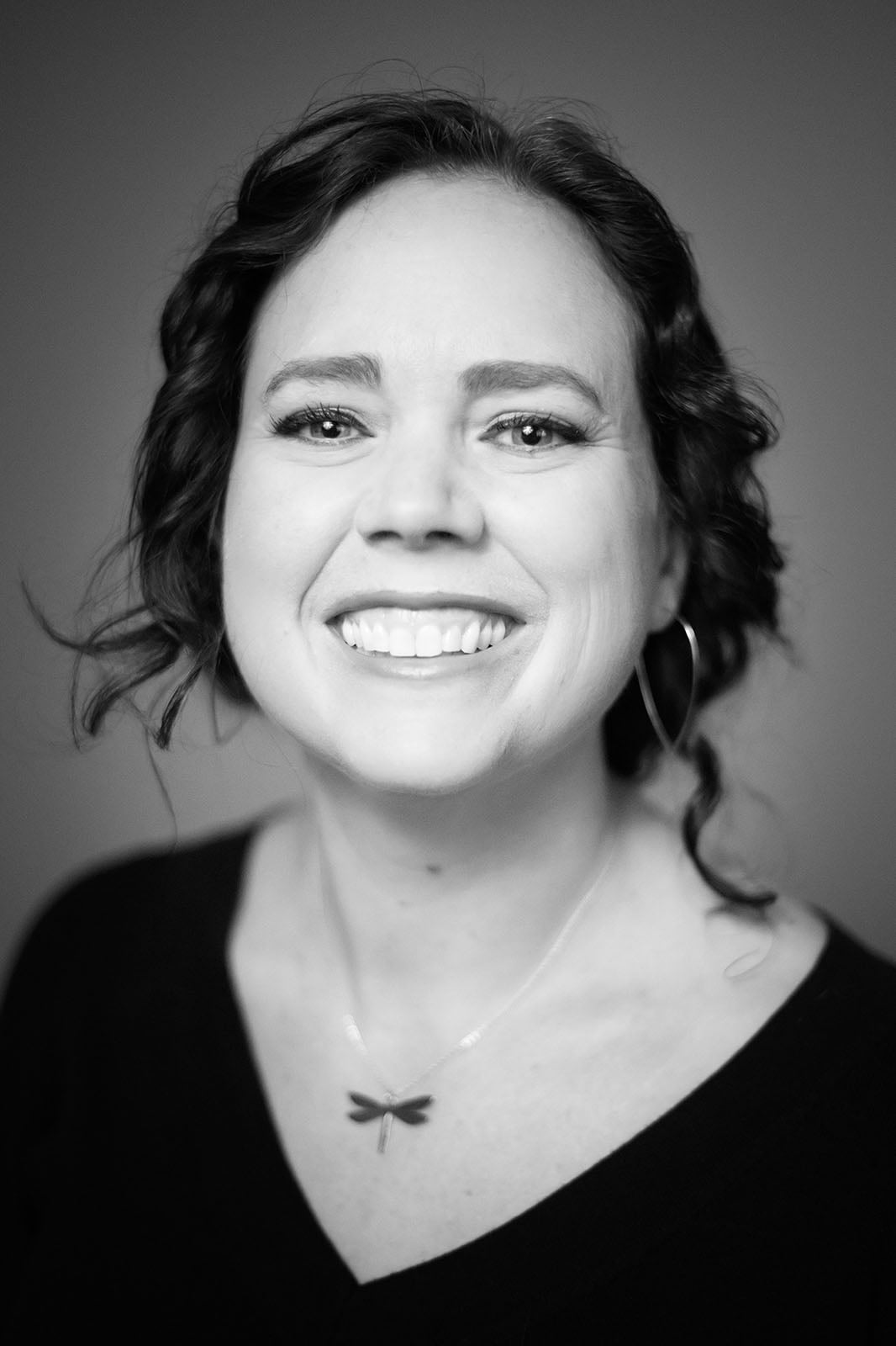 A woman with wavy dark hair smiles at the camera. She wears a dark top, hoop earrings, and a dragonfly necklace. The photo is in black and white, with a blurred background.
