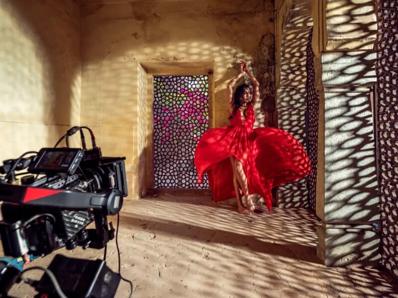 A woman in a flowing red dress dances in sunlight, casting intricate shadows on the walls, while a camera films her in a rustic room with a decorative screen.