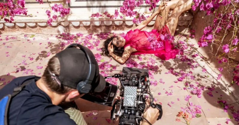 A photographer films a woman in a bright pink dress lying on the ground surrounded by pink flower petals, striking a pose against a decorative stone wall.