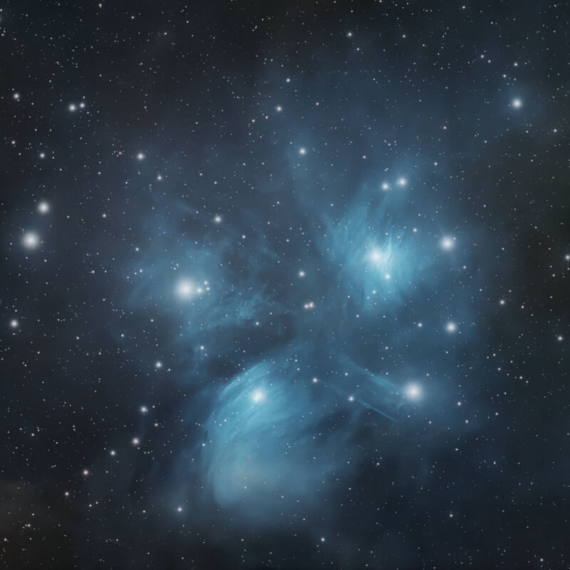 The Pleiades star cluster features bright, blue stars surrounded by wispy, glowing reflection nebulae against a dark, star-filled sky.