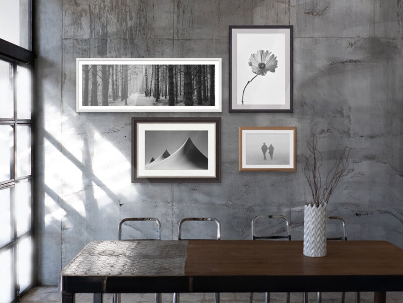 Industrial-style dining area with a wooden table, metal chairs, and gray concrete wall. Four framed black-and-white photos hang on the wall; a vase with twigs sits on the table. Sunlight casts shadows inside.