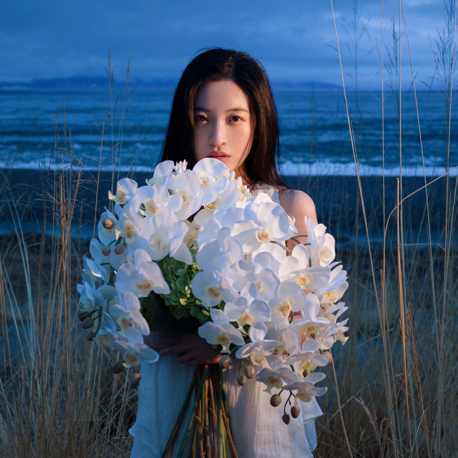 A young woman with long dark hair holds a large bouquet of white orchids while standing in tall grass by the ocean at dusk.