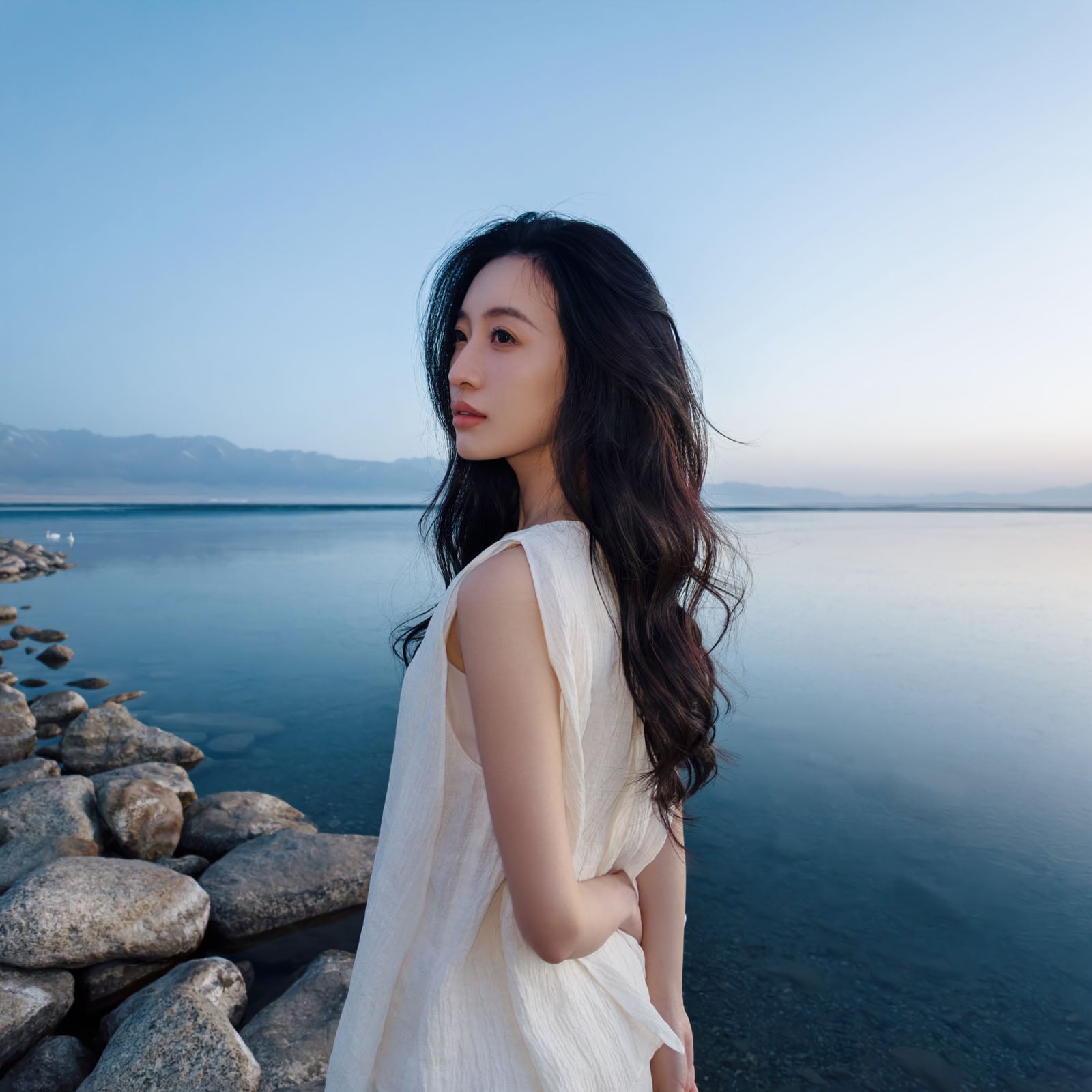 A woman with long, dark wavy hair wearing a white sleeveless dress stands by a rocky lakeshore, looking out toward distant mountains under a clear sky.