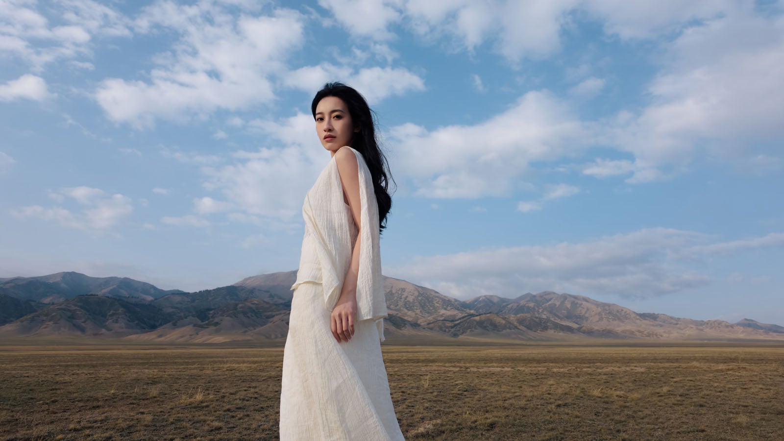 A woman with long dark hair wearing a white sleeveless dress stands in a vast, dry field with mountains under a blue sky.