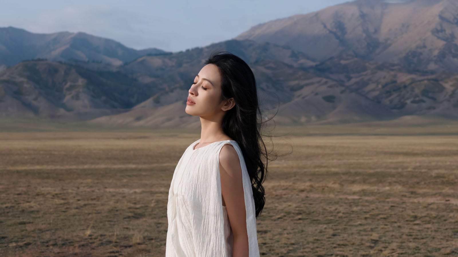 A woman with long dark hair wearing a white dress stands with her eyes closed in a vast, dry landscape with mountains.
