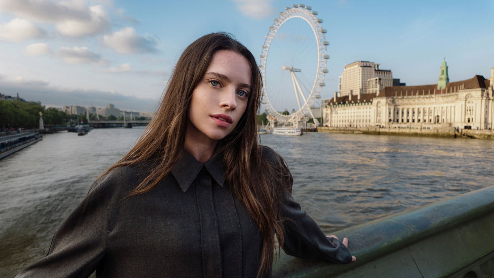 A woman with long brown hair stands on a bridge with the London Eye and a river in the background.