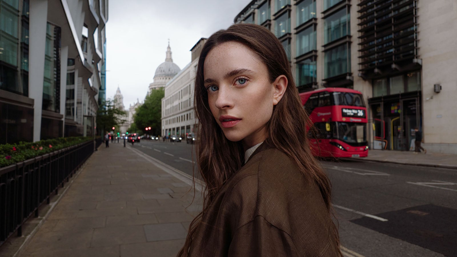 A young woman with long brown hair and freckles looks over her shoulder on a city sidewalk with a red bus and a dome in the background.