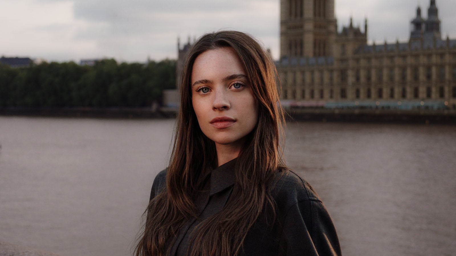 A young woman with long brown hair and freckles looks directly at the camera with a large stone building in the background.
