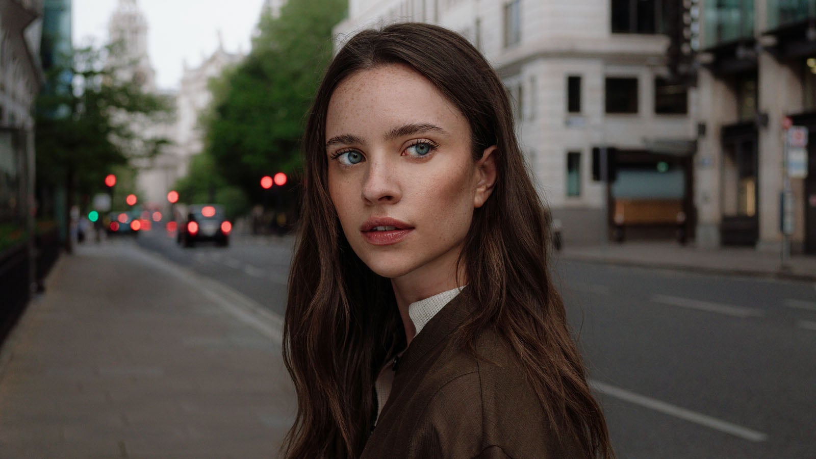 A young woman with long brown hair and freckles looks toward the side while standing on a city street.