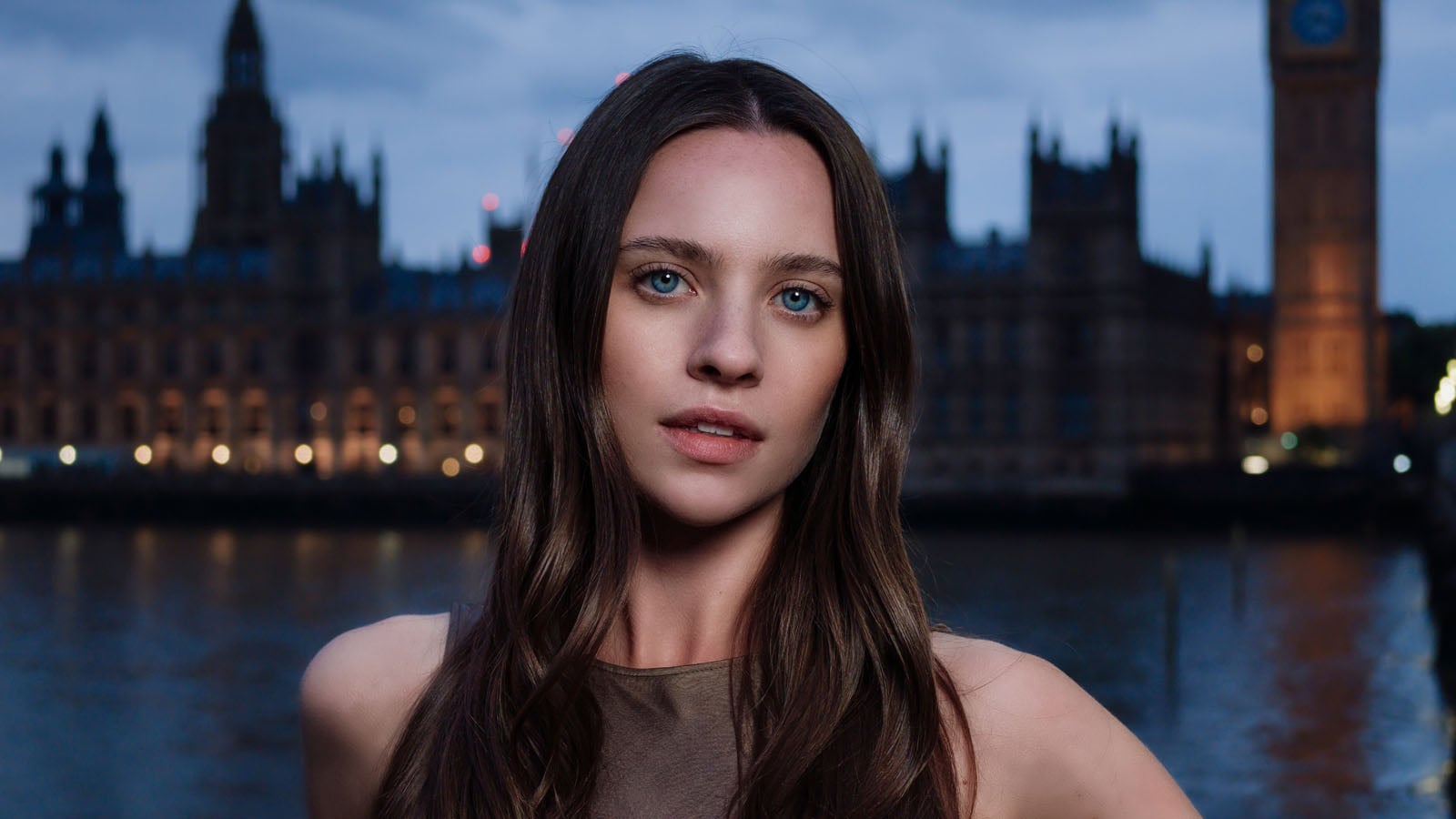 A young woman with long brown hair and blue eyes looks directly at the camera against a blurred backdrop of a city at dusk.