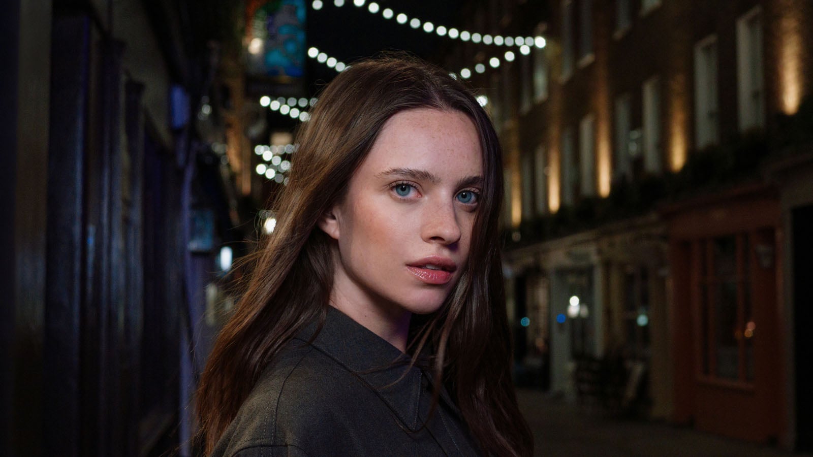 A young woman with long brown hair and blue eyes looks directly at the camera on a city street at night.