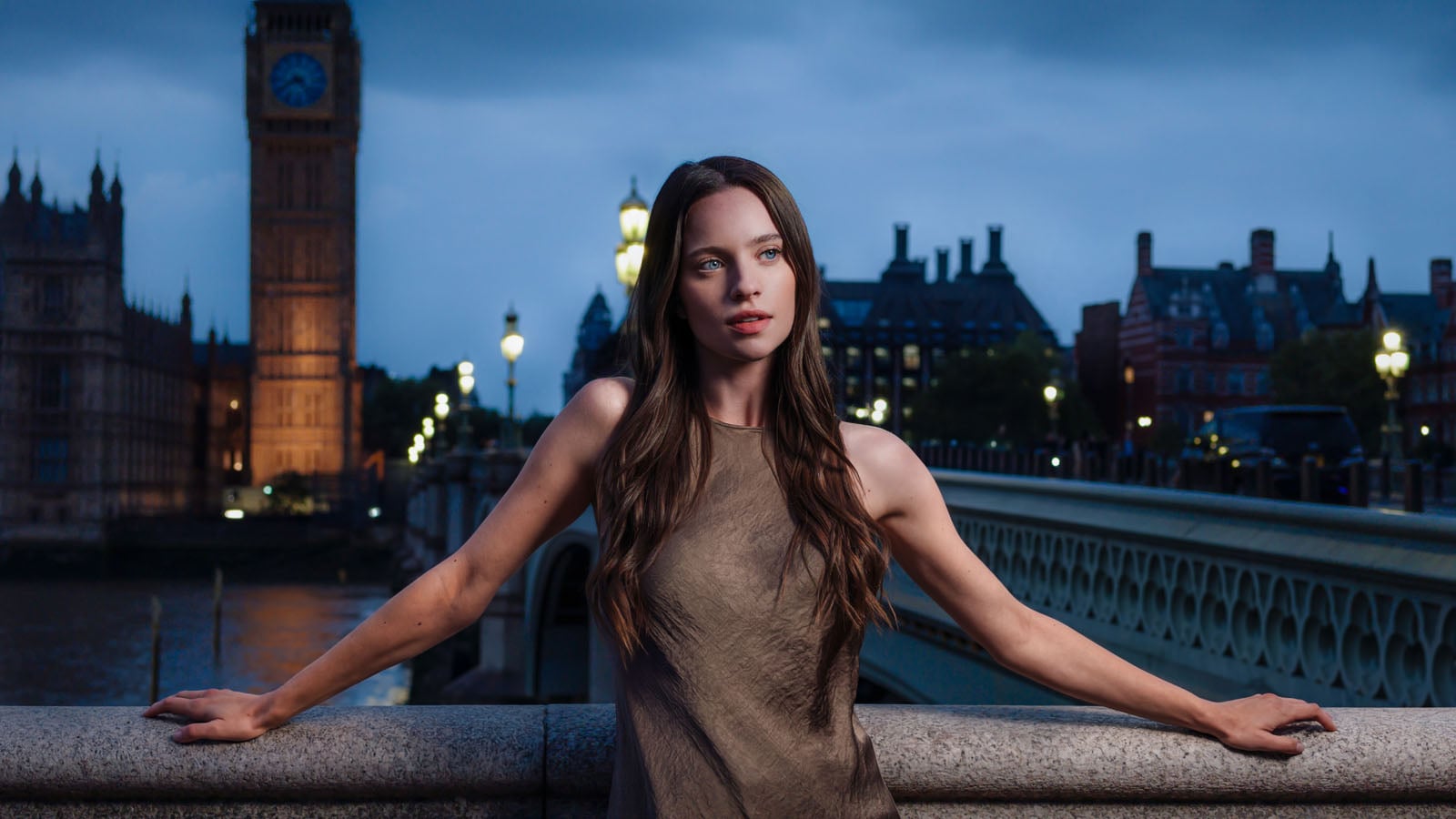 A woman with long brown hair stands on a stone bridge at dusk, with a large clock tower and city buildings in the background.