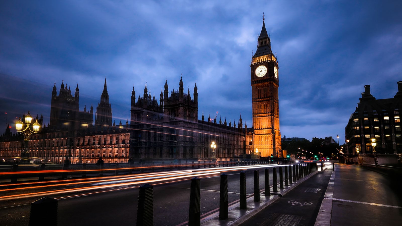 A long-exposure shot of the Elizabeth Tower and the Palace of Westminster at dusk, with light trails from passing traffic on the road in the foreground.